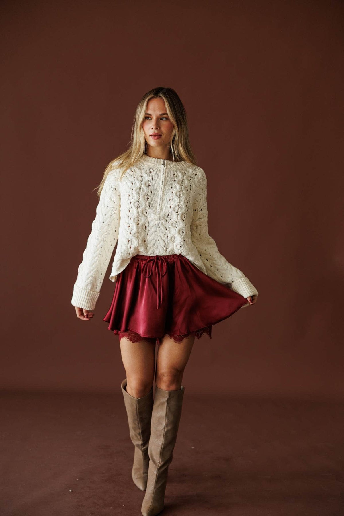 Cable Me Later Sweater in cream, full-length view styled with a burgundy satin skirt and tall boots